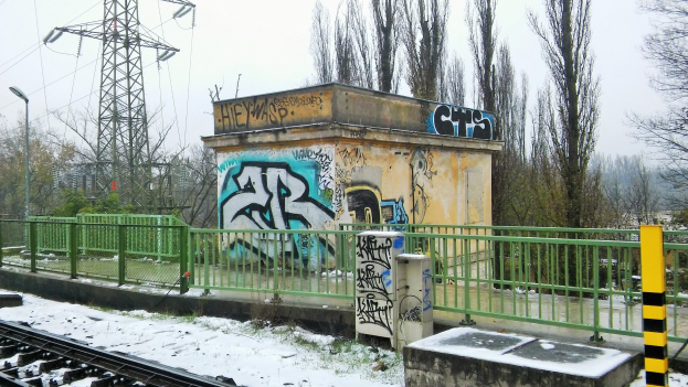 Eine Bahnstrecke mit Graffiti an der Seite, umgeben von Gelöndern, Pfosten, Lichtern und Bäumen, mit Törmen und Dröhnen im Hintergrund und Schnee am Boden.