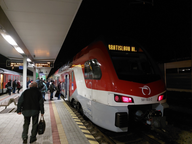 Roter und weißer Zug hält nachts auf einem Bahnhof mit Wartenden auf dem Bahnsteig und Bänken auf der linken Seite sowie Deckenbeleuchtung