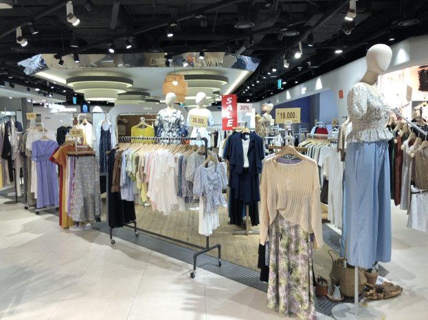 Bekleidungsgeschäftsauslage mit Schaufensterpuppen in Kleidern an Kleiderbügeln, beleuchtet von Deckenlampen und Werbetafeln mit Text.