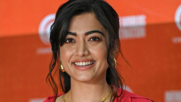 Eine Frau in einem roten Kleid mit einem eleganten Dutt und dezentem Make-up lächelt in die Kamera, trägt schwarze High Heels und hat ein hellgelbes Banner im Hintergrund mit der Aufschrift "Sonam Kapoors schönste Looks vom Cannes Film Festival".