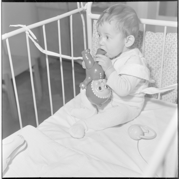 Ein Schwarz-Weiß-Foto eines Babys in einer Wiege, das ein Spielzeug hält und es ansieht, umgeben von einer Wand.