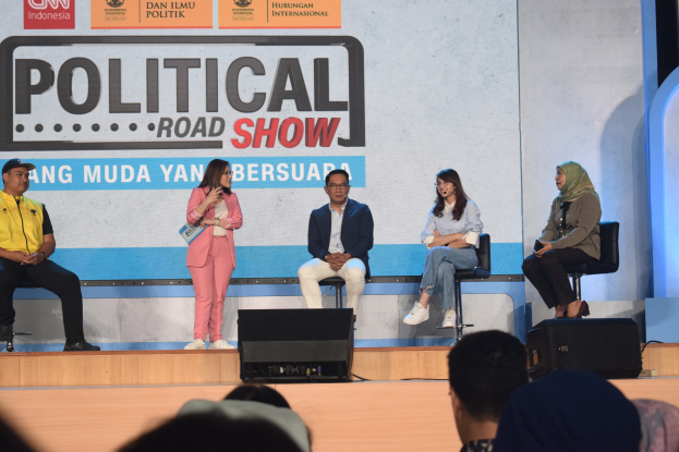 Eine Gruppe von Menschen sitzt auf einer Bühne mit einer Frau in der Mitte, die ein Mikrofon hält, einem Publikum im Vordergrund und einem Banner mit der Aufschrift 'Politischer Straßenumzug 2019' im Hintergrund.