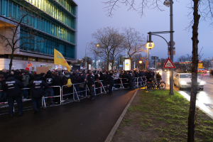 Eine große Gruppe von Menschen steht vor einem Gebäude in Berlin, hält Schilder, mit Barrikaden, Fahrrädern, Laternen, Schildern, Bäumen und Gras auf dem Boden, unter einem sichtbaren Himmel.