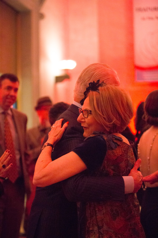Ein Mann und eine Frau umarmen sich herzlich auf einer Party, umgeben von anderen Menschen mit einer beleuchteten Wand im Hintergrund.