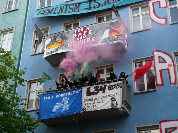 Eine Gruppe von Menschen auf einem Balkon mit Fahnen und Transparenten, links ein Baum und Rauch in der Luft, was auf eine Demonstration hindeutet.