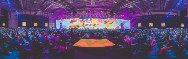 Eine große Menschenmenge sitzt auf Stühlen und schaut auf eine hell erleuchtete Bühne mit mehreren stehenden Personen, wahrscheinlich auf einem Online-Marketing-Rockstars-Event, umgeben von Bildschirmen.