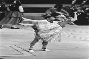 Ein Schwarz-Weiß-Foto eines tanzenden Paares auf einer Bühne, mit einigen Menschen auf der Treppe im Hintergrund und einem Pfahl im Vordergrund.