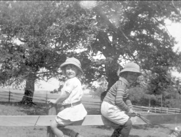 Zwei junge Jungs in Mützen halten Baseballschläger in einem grünen Park mit Bäumen und einem Zaun im Hintergrund, die Szene in Schwarz-Weiß.
