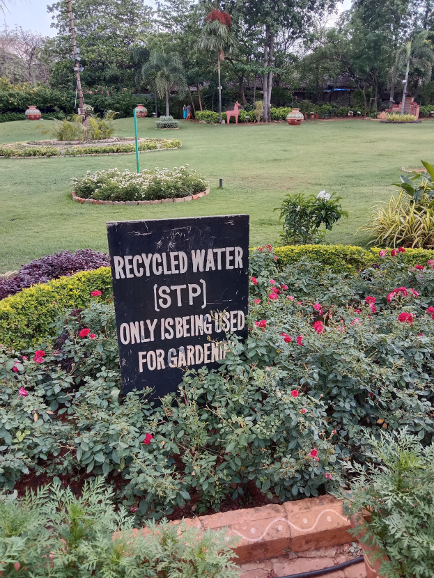 Schilder mit der Aufschrift "Recycled Water (STP) Only Being Used for Gardening" in einem Garten mit saftig grünem Gras, leuchtenden Blumen, hohen Bäumen und einem hellblauen Himmel.