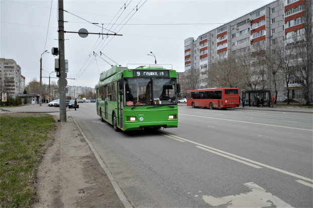 Ein grüner Bus fährt eine Straße entlang, die von hohen Gebäuden gesäumt ist, mit Gras und Strommasten auf der linken Seite, Bäumen im Hintergrund und einem klaren blauen Himmel darüber.