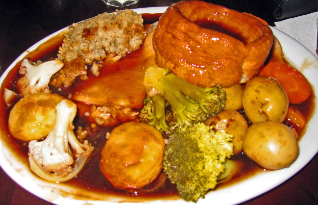 Ein weißer Teller mit Yorkshire-Pudding und einer Auswahl an Gemüsen wie Karotten, Kartoffeln und Zwiebeln, die auf einem Tisch mit Gläsern, Papiertüchern und anderen Gegenständen drumherum stehen.