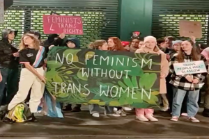 Eine Gruppe von Menschen, die auf der Straße stehen und ein Banner mit der Aufschrift "Kein Feminismus ohne Trans-Frauen" sowie verschiedene Schilder halten, mit einer Wand und einem Rollladen im Hintergrund.
