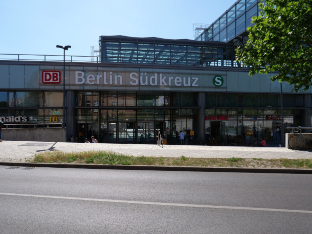 Ein großes modernes Bahnhofsgebäude mit Glaswänden und Text, umgeben von Straßeninfrastruktur, Fahrzeugen, Fußgängern, Fahrrädern, Bäumen und einem klaren blauen Himmel.
