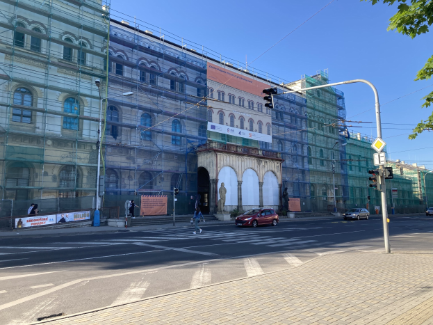 Großes Gebäude mit Gerüst während der Renovierung umgeben von Fahrzeugen, Passanten, Ampeln, Straßenschildern, Pfosten, Bäumen und einem klaren blauen Himmel.