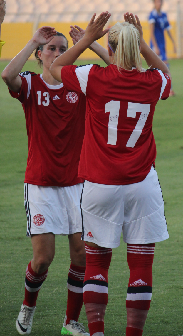 Zwei Frauen in roter T-Shirts und weißen Shorts stehen auf einem Fußballfeld und klatschen in die Hände, während im Hintergrund eine Person in einem blauen Kleid, eine gelbe Tafel und einige Stühle zu sehen sind.