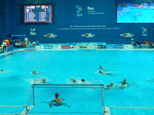Eine Gruppe von Menschen spielt Wasserball in einem Pool während der FINA-Weltmeisterschaften 2016 in Kazan, mit einem Netz, das den Pool trennt, und Zuschauern auf der linken Seite.
