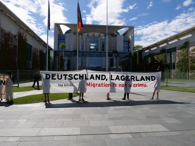 Eine Gruppe von Menschen hält ein Banner mit der Aufschrift "Deutschland, Lagerland Migration ist kein Verbrechen" vor einem Gebäude mit Fenstern, umgeben von Gras, einem Metallzaun, Pflanzen, Bäumen und Fahnenstangen unter einem bewölkten Himmel.