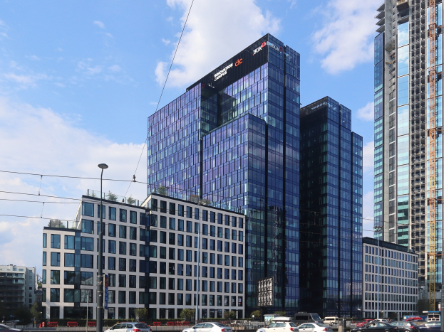 Großes modernes Bürogebäude mit zahlreichen Fenstern, identifiziert als die Deutsche Bank Hauptverwaltung in Frankfurt, Deutschland, umgeben von städtischer Infrastruktur einschließlich Straßenlaternen, Fahrzeugen, Grünflächen und einem bewölkten Himmel.