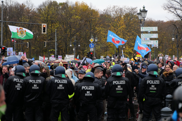 Eine große Gruppe von Polizisten steht vor einer Menge mit Fahnen und Schirmen, im Hintergrund sind Laternenpfähle, Verkehrsampeln, Schilder, Bäume und ein klarer Himmel zu sehen.