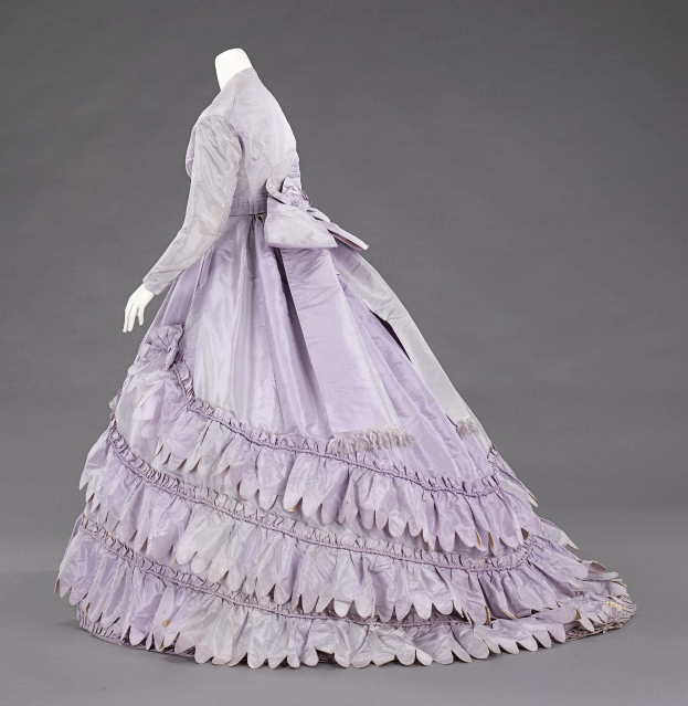 Eine Schaufensterpuppe in einem lila Ballkleid mit Rüschen an den langen Ärmeln, einem eng anliegenden Oberteil und einem bodenlangen Rock vor einem grauen Hintergrund.