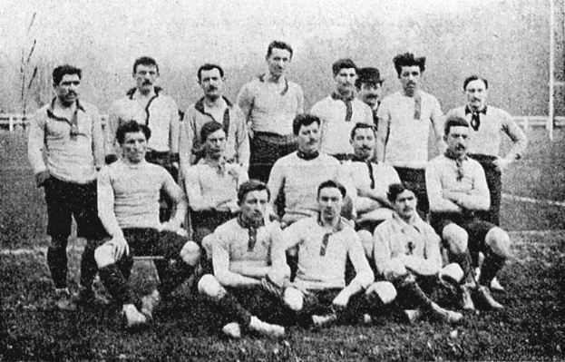 Ein Schwarz-Weiß-Foto einer Fußballmannschaft aus den 1920er Jahren, mit einigen stehenden und anderen sitzend auf dem Boden, vor Pfählen, Bäumen und einem klaren Himmel.