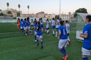 Eine Gruppe von Frauen, die Fußball auf einem Außenfeld spielen, mit Gebäuden, Bäumen und einem Tor im Hintergrund.