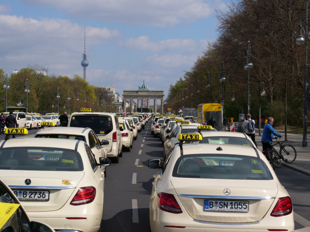 Eine lange Reihe von Taxis, die an einer belebten Straße in Berlin, Deutschland, geparkt sind, mit fahrenden Fahrzeugen, Radfahrern, Fußgängern, Bäumen, Laternenmasten, Gebäuden, einem Bogen und einem Turm im Hintergrund unter einem bewölkten Himmel.