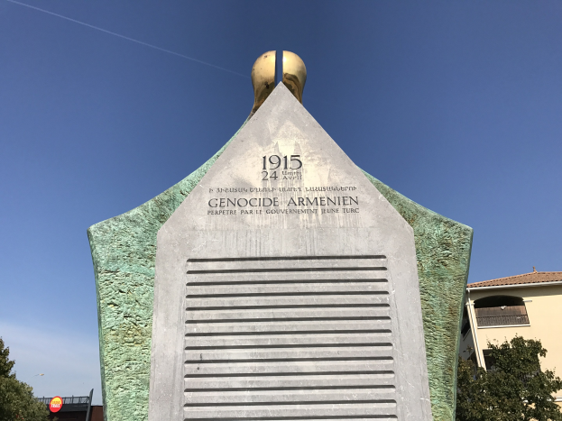Ein hoher Denkmal mit einer Uhr darauf, mit Inschrift, steht in einem Park umgeben von Bäumen und Gebäuden unter einem klaren Himmel, zum Gedenken an den 24. Jahrestag des Völkermords in Armenien.