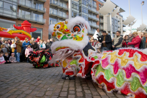 Eine Löwen-Tanzvorstellung während einer Chinesischen Neujahrsfeier in Amsterdam, mit einer Zuschauermenge und Menschen, die den Moment mit Kameras festhalten, vor einer Kulisse aus Gebäuden und einem klaren blauen Himmel.