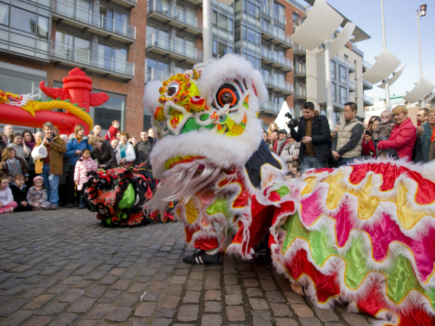 Eine Löwen-Tanzvorstellung während einer Chinesischen Neujahrsfeier in Amsterdam, mit einer Zuschauermenge und Menschen, die den Moment mit Kameras festhalten, vor einer Kulisse aus Gebäuden und einem klaren blauen Himmel.