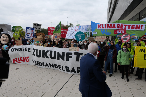 Eine Gruppe von Menschen, die gegen den Klimawandel protestieren und Schilder und Banner in der Hand halten, vor einem Gebäude stehend, mit einer Person, die eine Kamera hält, auf der linken Seite und Bäumen, Pfählen und einem klaren blauen Himmel im Hintergrund.
