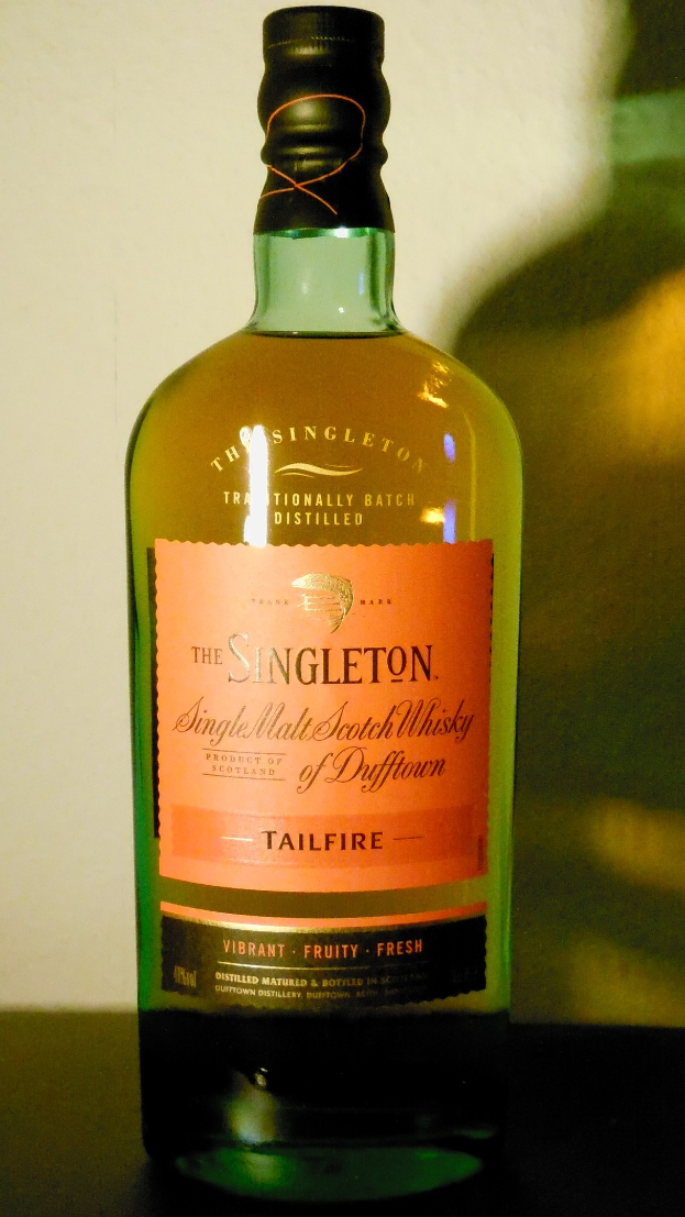 Eine tiefe bernsteinfarbene Flasche Singleton of Dufftown Tailfire Single Malt Scotch Whisky auf einem Tisch, mit einem Etikett, das eine Abbildung mit einem Löwen und einem Schloss enthält.