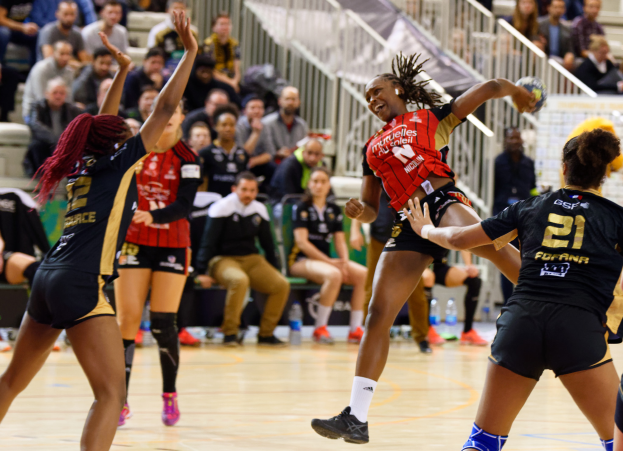 Eine Gruppe von Frauen spielt Volleyball auf einem Platz mit einem Ball in der Mitte, während Zuschauer auf Stühlen und Geländern im Hintergrund sitzen und Flaschen auf dem Boden liegen.