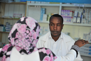 Eine Apothekerin in einem weißen Kittel spricht mit einer sitzenden Frau in einer Apotheke, mit Regalen voller Schachteln und anderer Gegenstände dahinter.