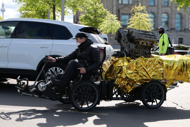 Ein Mann im Rollstuhl mit einem großen Motor auf dem Rücken, umgeben von Fahrzeugen auf einer Straße mit Bäumen, Gebäuden, Polen und einem klaren blauen Himmel im Hintergrund, trägt eine schwarze Jacke und eine Mütze, während er ein Objekt hält.
