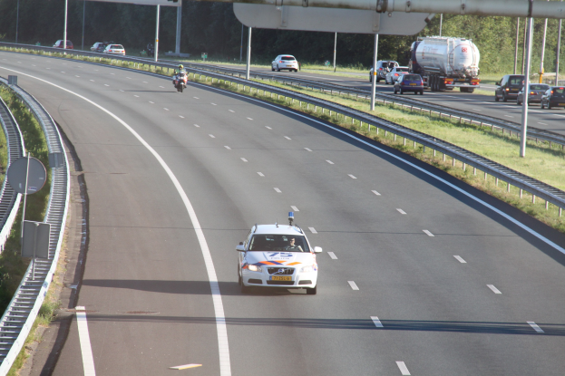Ein Auto auf einer Autobahn mit einem stationären Polizeiwagen am Rande, andere Fahrzeuge und Baumbestände im Hintergrund.
