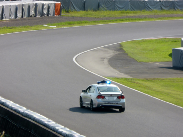 Polizeifahrzeug auf einer Rennstrecke mit Gras auf beiden Seiten, Menschen und Zelte im Hintergrund.