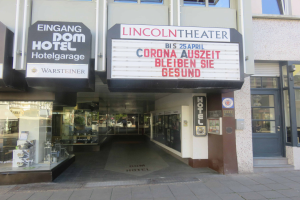 Das Lincoln Theater in Berlin, Deutschland, ein Gebäude mit Glasfenstern und -türen und einer Tafel mit Text darauf, mit verschiedenen Objekten im Inneren, die den Eindruck einer pulsierenden Stadtlandschaft vermitteln.
