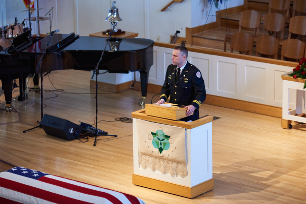 Ein Polizist in Uniform steht an einem Podium mit einem Mikrofon und spricht vor einem Publikum in einem Raum mit einem Piano, Musikinstrumenten und diverser Ausrüstung im Hintergrund.