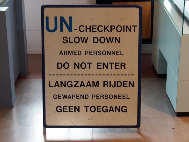 Schild auf dem Boden mit der Aufschrift "UN-Checkpoint Verlangsame bewaffnete Personen - Betreten verboten" mit einer Glasplatte und Gegenständen rechts und einer Wand mit einem Gegenstand im Hintergrund.