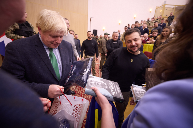 Mann in Anzug und Krawatte signiert Autogramme für eine Menschenmenge, einige halten Taschen und Bücher, mit einer beleuchteten Wand im Hintergrund.
