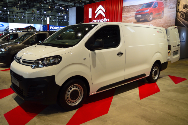 Ein weißer Citroen Berlingo Van wird auf einer Automesse präsentiert, umgeben von schwarzer Schlafzimmer-Möblierung, während die Wände mit Brettern und Lichtern geschmückt sind.