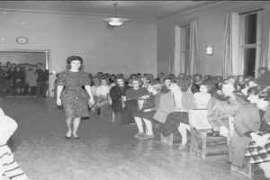 Eine Frau in einem schwarzen und weißen Kleid läuft über den Laufsteg während einer Mode-Show für Frauenrechte, mit einer sitzenden Menge und einer Uhr und Lampen an der umliegenden Wand.