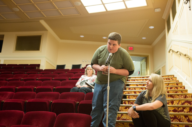 Ein Mann in einem grünen T-Shirt und blauen Jeans steht vor einem Publikum in einem Theater, hält ein Mikrofon in der Hand, mit zwei Frauen auf der Treppe dahinter, roten Stühlen im Hintergrund, einem Geländer auf der rechten Seite und Lampen auf dem Dach.