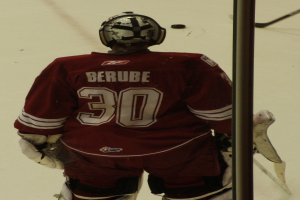 Ein Hockey-Spieler steht auf dem Eis vor einem Tor, trägt einen Helm, Handschuhe und ein Jersey mit Text und Nummern, mit einem Pfosten rechts daneben.