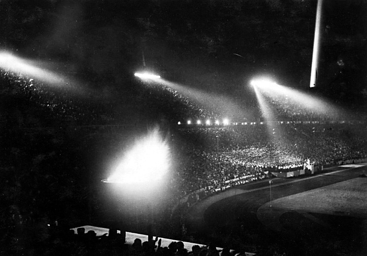 Schwarzes und weißes Foto eines vollen Stadions unter Flutlicht vor dunklem Hintergrund.