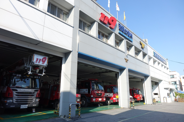 Feuerwache mit mehreren Feuerwehrautos vor dem Gebäude, mit Fenstern, Säulen, Namensschildern und Flaggen im Vordergrund; andere Gebäude und ein klarer blauer Himmel im Hintergrund.