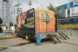 Ein Lieferwagen parkt vor einem Gebäude mit einer Tafel, auf der "Teilen Sie eine positiv überladene Erfahrung" steht, mit Stufen, einem Zylinder, Geländern, Strommasten, Laternen, Bäumen und einem bewölkten Himmel.