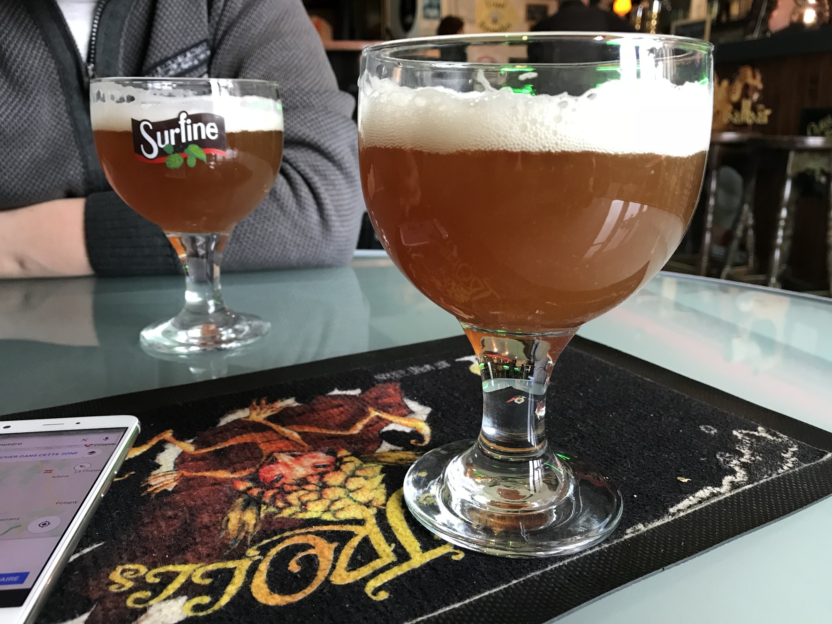 Zwei Gläser goldenes Bier auf einem Tisch neben einem Handy, mit Menschen und gerahmter Wanddekoration im Hintergrund.