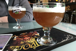 Zwei Gläser goldenes Bier auf einem Tisch neben einem Handy, mit Menschen und gerahmter Wanddekoration im Hintergrund.